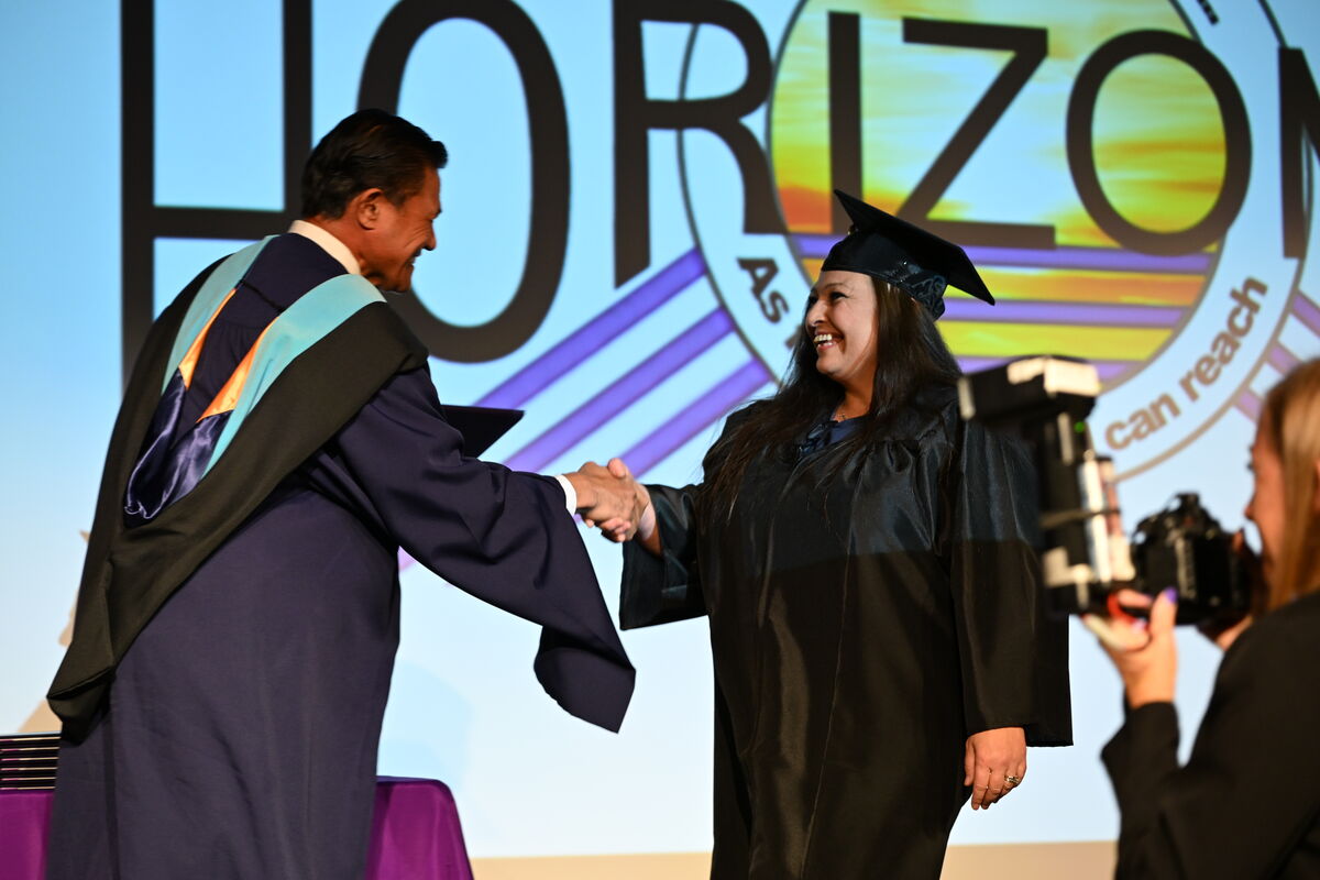 A smiling graduate in a cap and gown shakes hands with a faculty member on stage during a commencement ceremony.