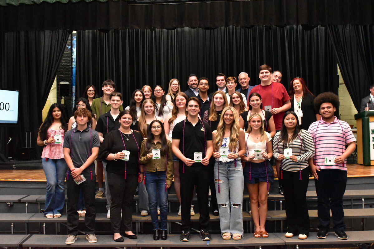 A diverse group of students and adults pose for a photo on tiered risers in front of a black curtain.
