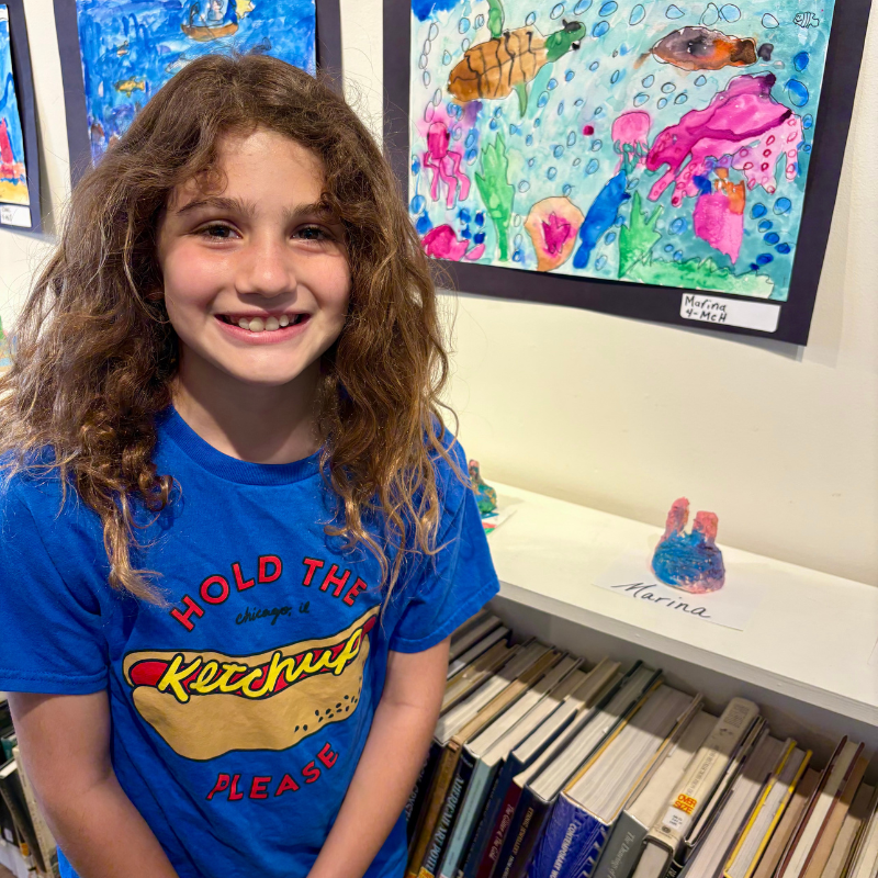 A young girl with curly hair smiles in front of artwork and a bookshelf.
