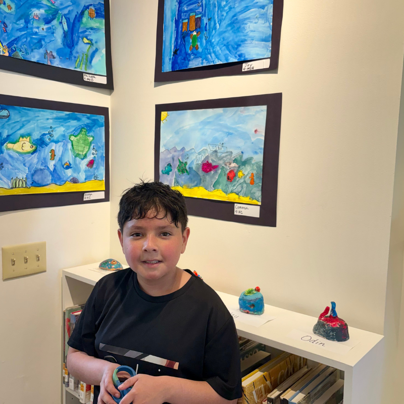 A young boy smiles in front of a wall displaying colorful children's artwork, including underwater scenes.