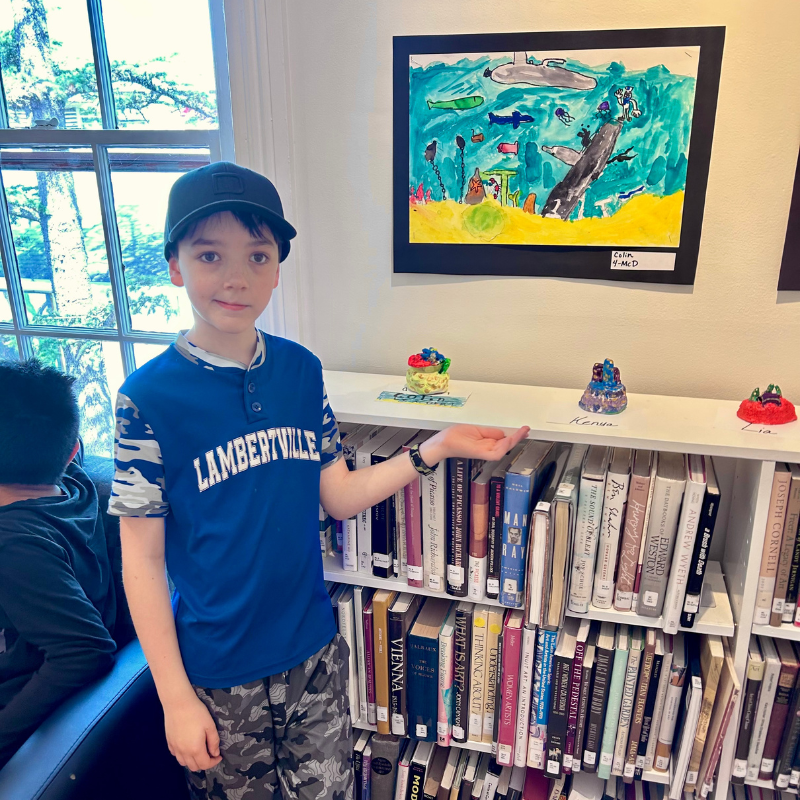 A young boy in a blue shirt stands next to a bookshelf displaying artwork and books.