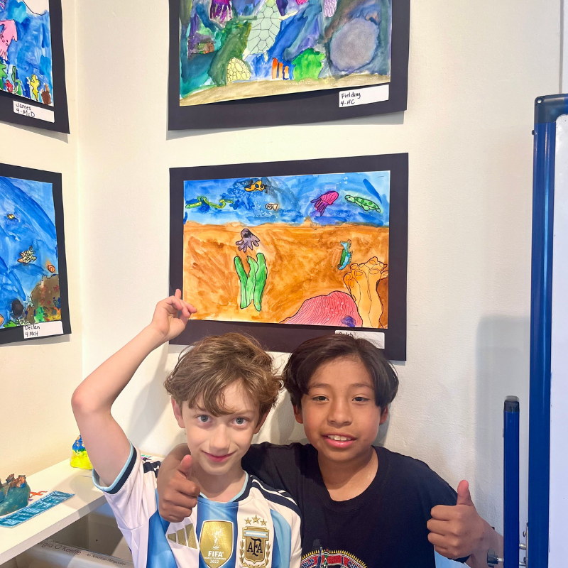 Two smiling boys give thumbs up in front of colorful underwater-themed artwork.