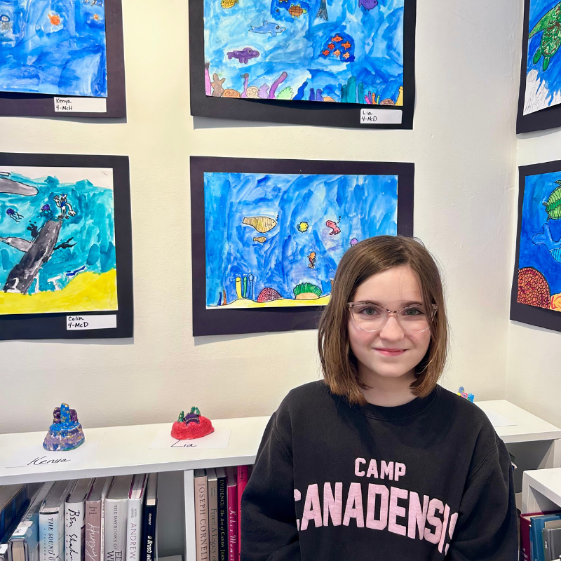 A young girl with glasses smiles in front of a wall displaying colorful children's artwork of underwater scenes.