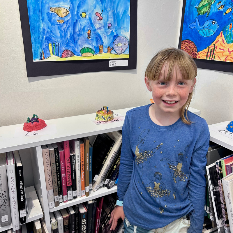 A young girl with blonde hair smiles in front of artwork and bookshelves.