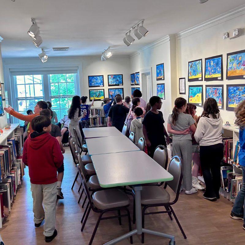 Children and adults observe artwork displayed on a wall in a room with bookshelves and tables.