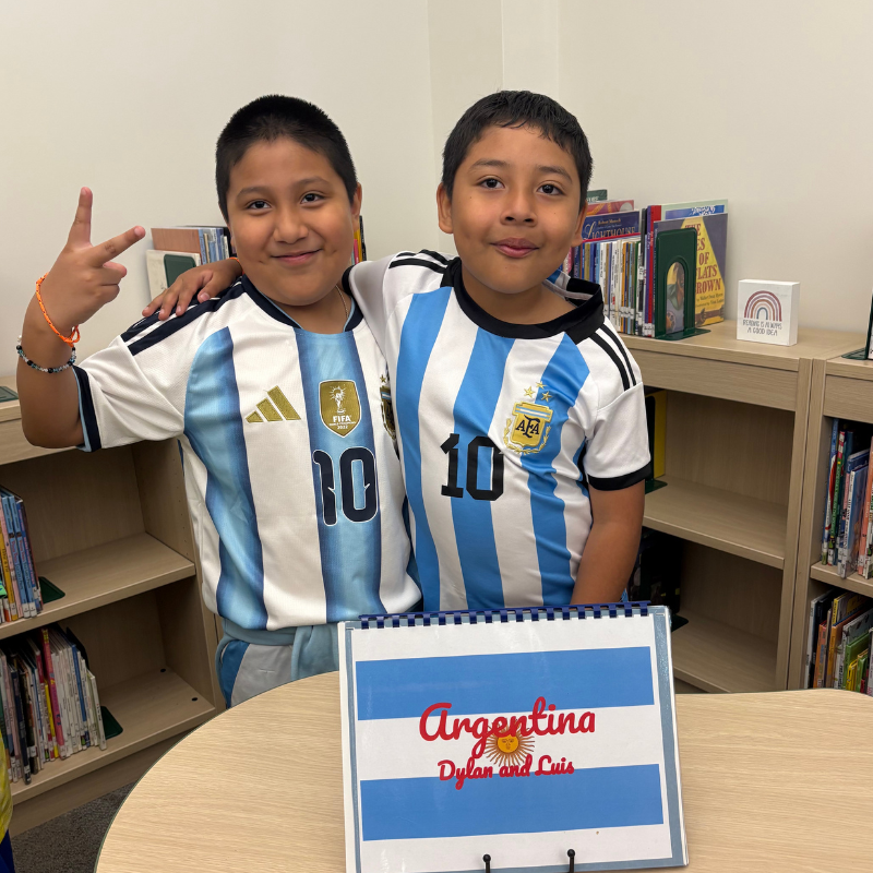 Two young boys in matching Argentina soccer jerseys stand together, one making a peace sign.