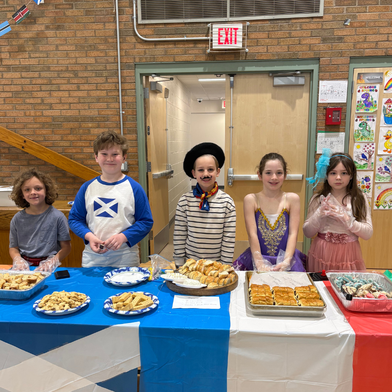 Four children stand behind a table displaying baked goods, with flags and artwork in the background.