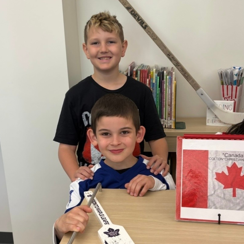 Two smiling boys stand behind a desk, one with hands on the other's shoulders.