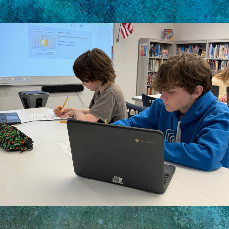 Two students focus on their work at desks in a classroom, one using a Chromebook and the other writing with a pencil.