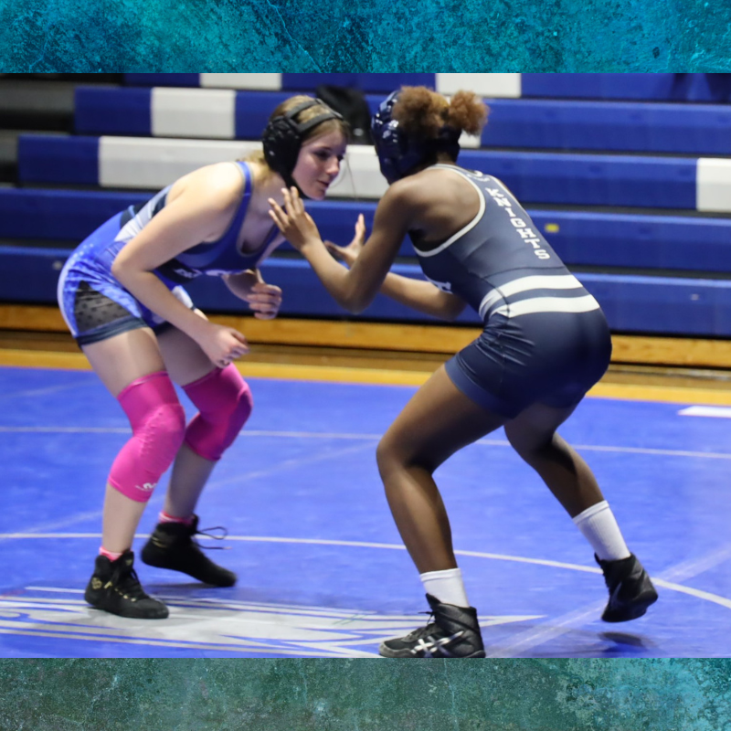 Two female wrestlers face off on a blue mat in a gymnasium.