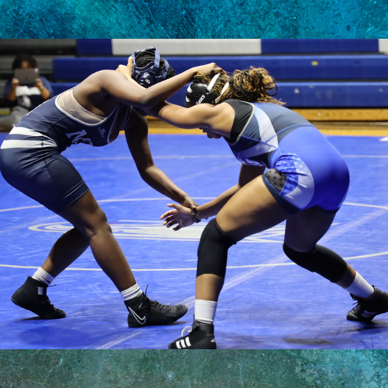 Two female wrestlers in blue and white uniforms grapple on a blue wrestling mat.