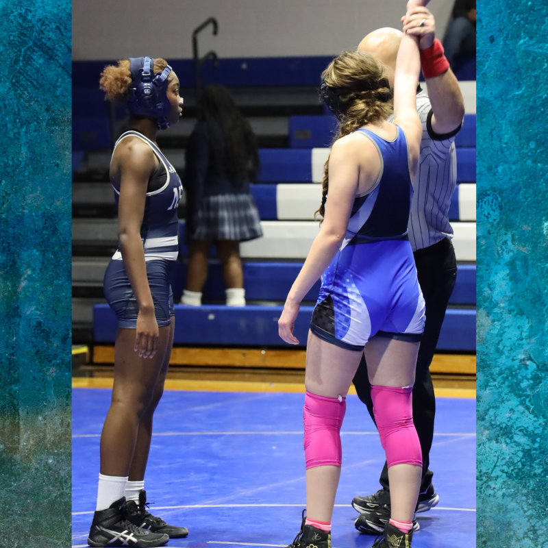 Two female wrestlers face each other on a blue mat, with a referee raising one wrestler's arm.