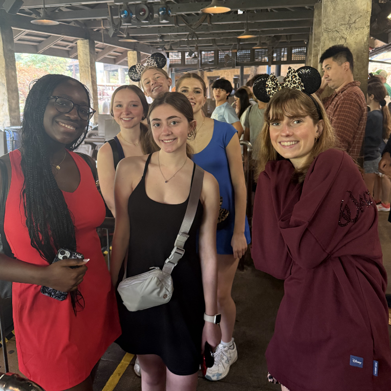 A group of young women wearing Mickey Mouse ears smiles while waiting in line.