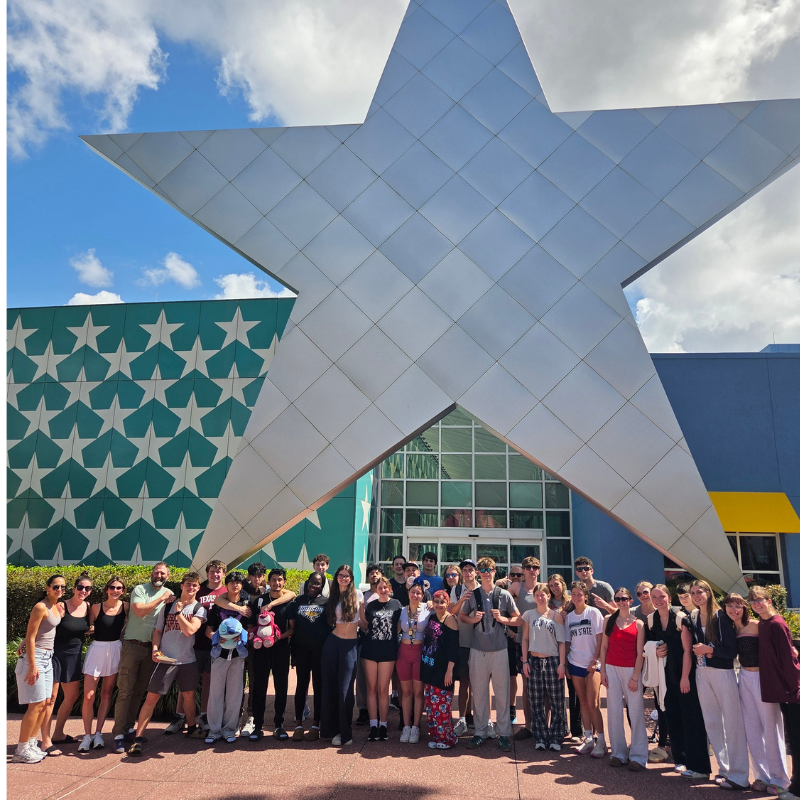 A large group of young people poses for a photo in front of a giant silver star and a teal building with white stars.