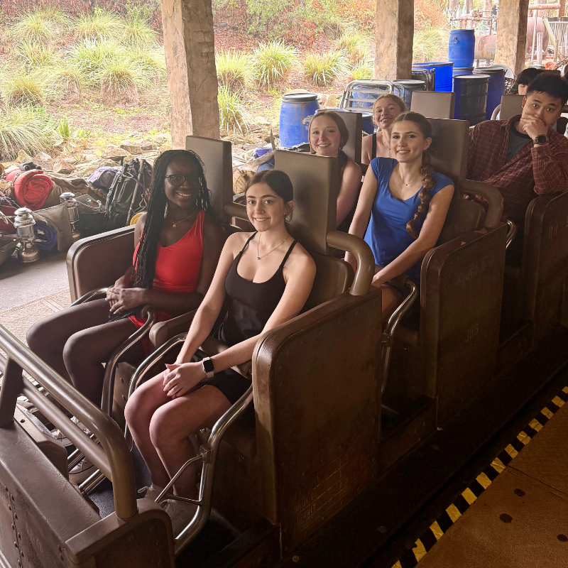 A group of young adults smiles while seated in a ride vehicle, anticipating an adventure.
