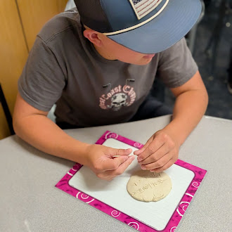 A person wearing a cap sits at a table and works with clay on a pink mat.
