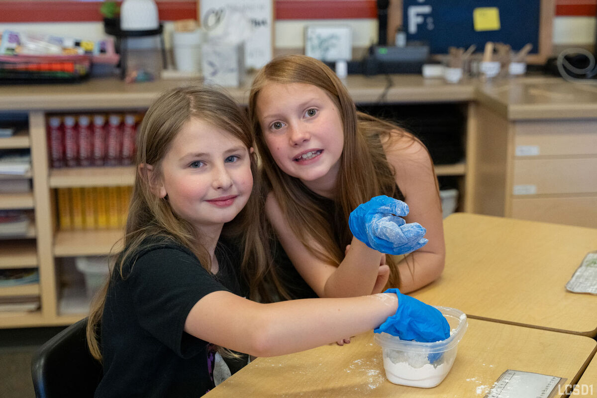 Two young girls wearing blue gloves work with white powder in a classroom setting.