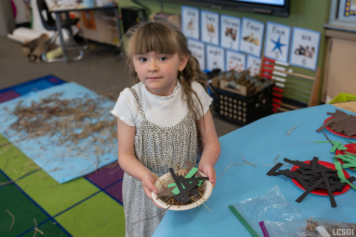 A young girl holds a small bowl filled with straw and craft materials, looking at the camera.