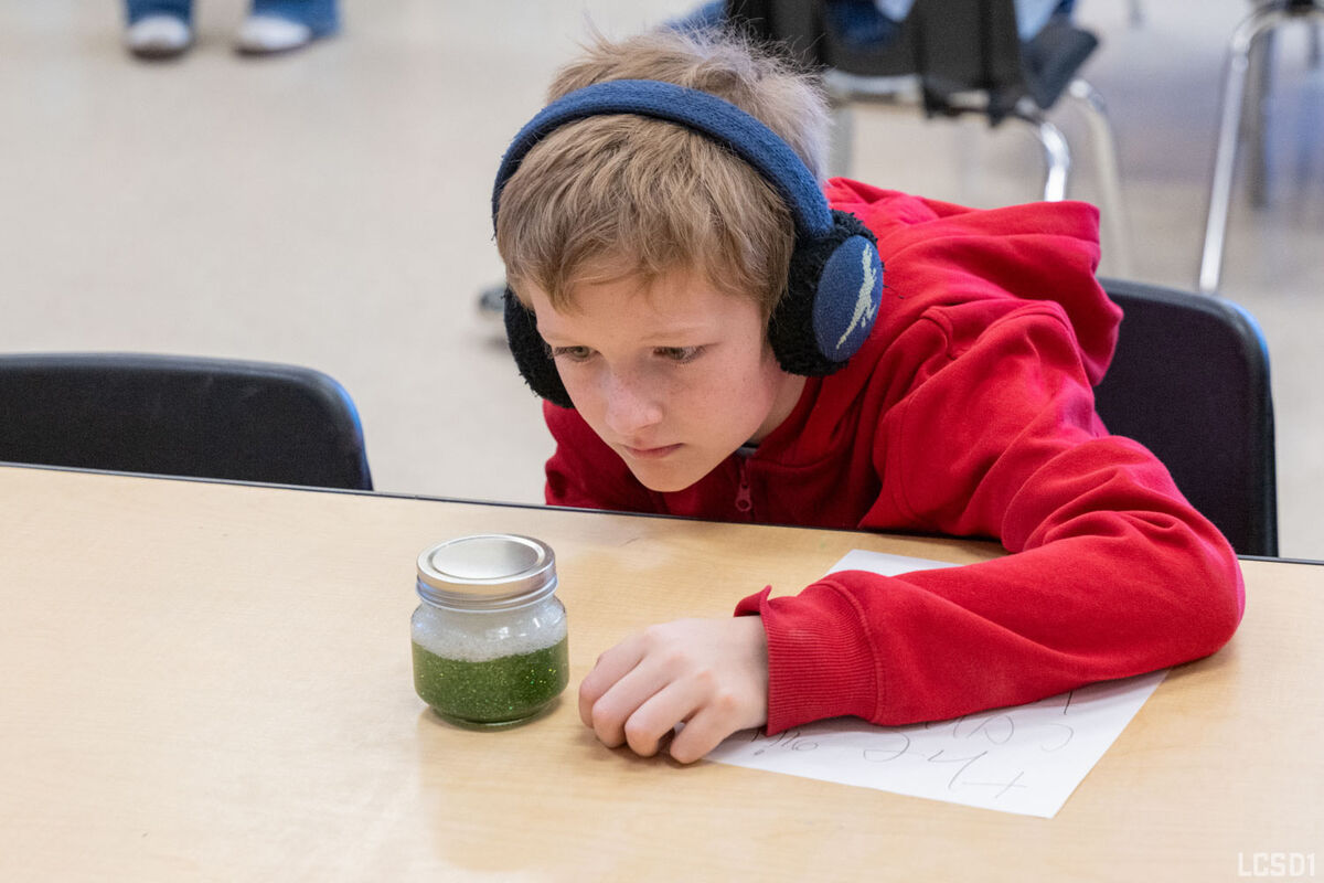 A young boy wearing headphones looks intently at a jar of green glitter and bubbles on a wooden table.