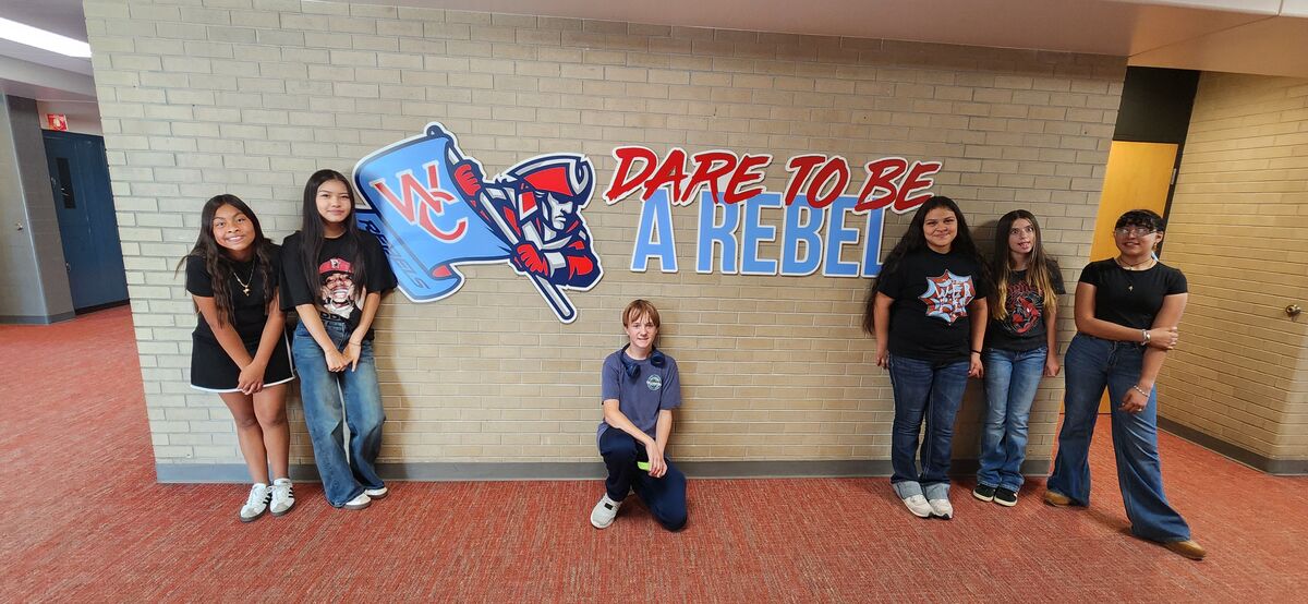 Six students pose together in front of a brick hallway wall featuring the school mascot and the text "Dare to be a Rebel".