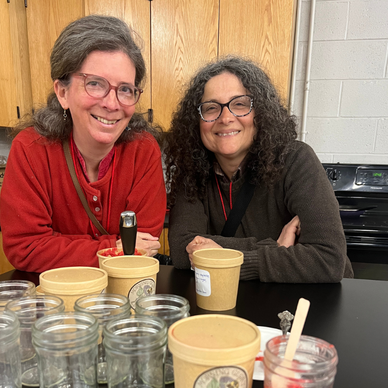 Two smiling women with glasses are seated at a table with jars and containers.
