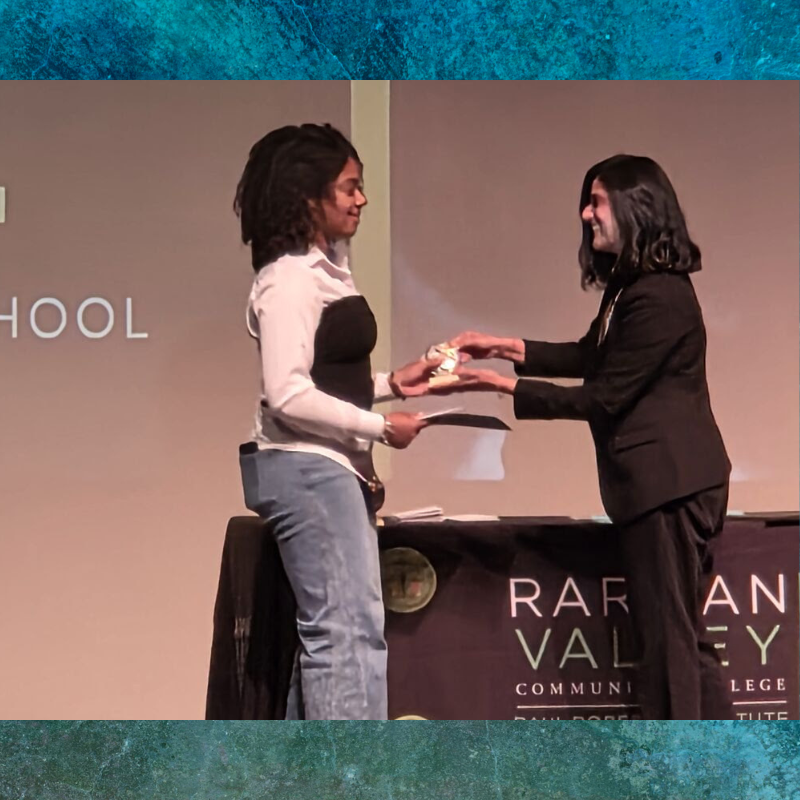 A student receives an award from an instructor on a stage, with 'HIGH SCHOOL' visible on the backdrop.