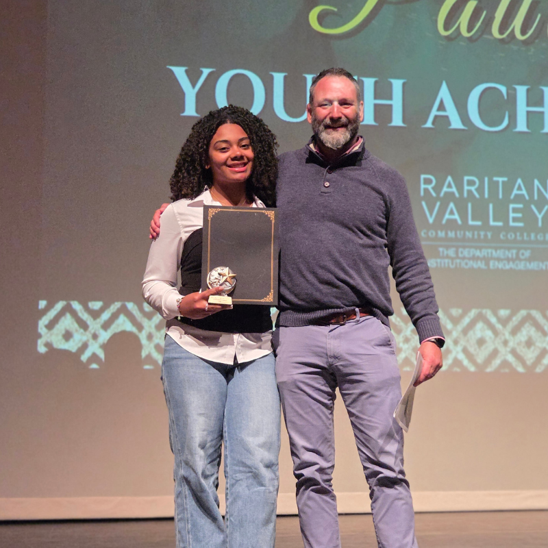 A young woman smiles as she holds a framed award and trophy, standing next to a man on a stage.