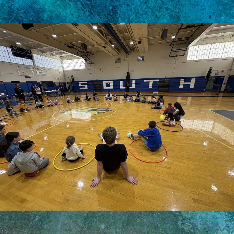 Children and adults sit in a circle on a gymnasium floor, surrounded by colorful hoops.
