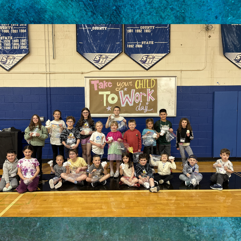 A group of children stand and sit on a gymnasium floor, holding bags and white objects, with a 'Take Your Child To Work Day' sign behind them.