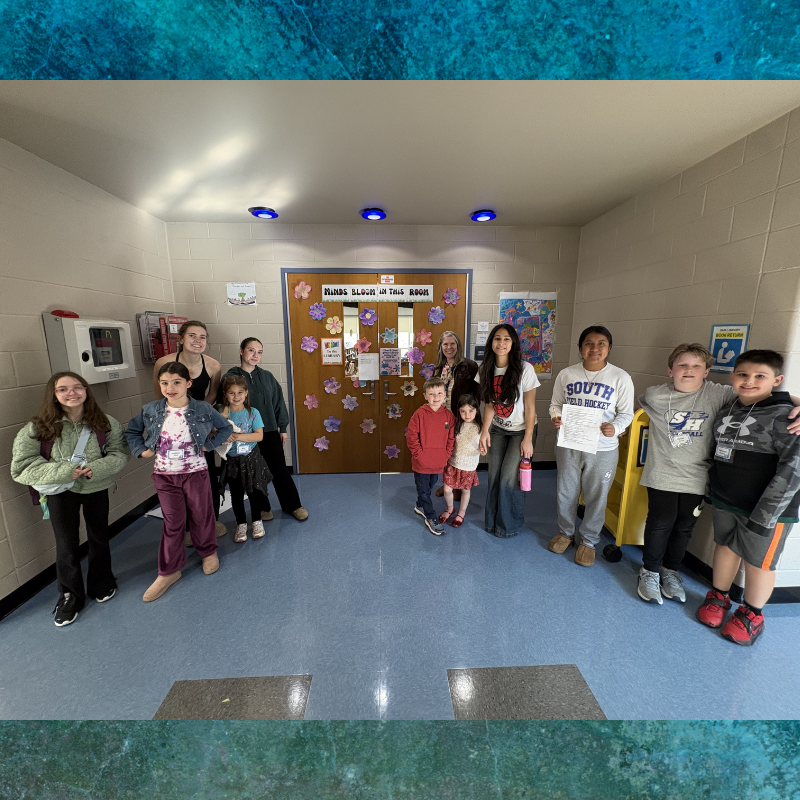 A group of children and adults stand in a hallway decorated with flowers and a sign that reads 'Minds Bloom in This Room'.
