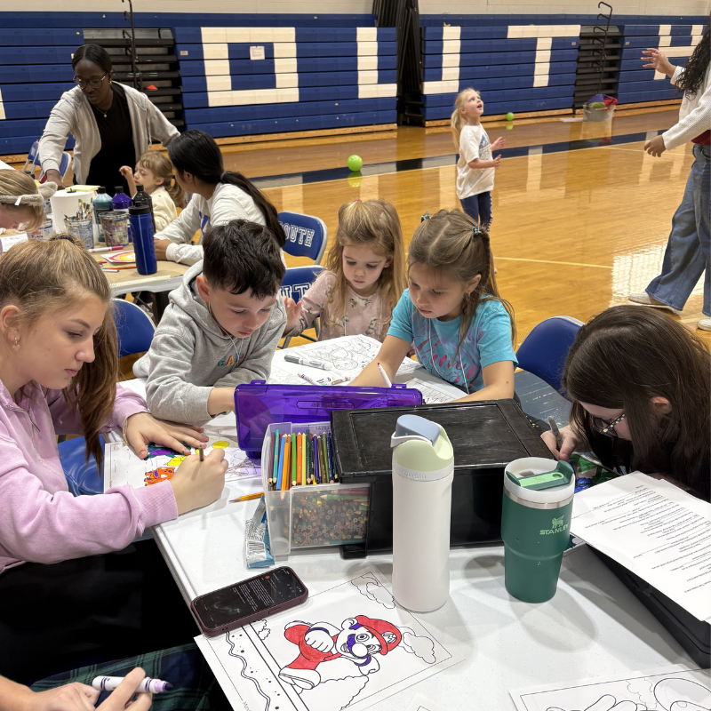 Children and adults engage in a coloring activity at tables in a gymnasium.