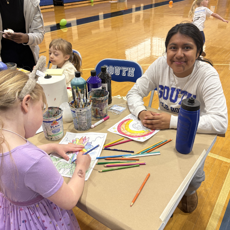 Children and a young woman are seated at a table, coloring pictures with pencils.