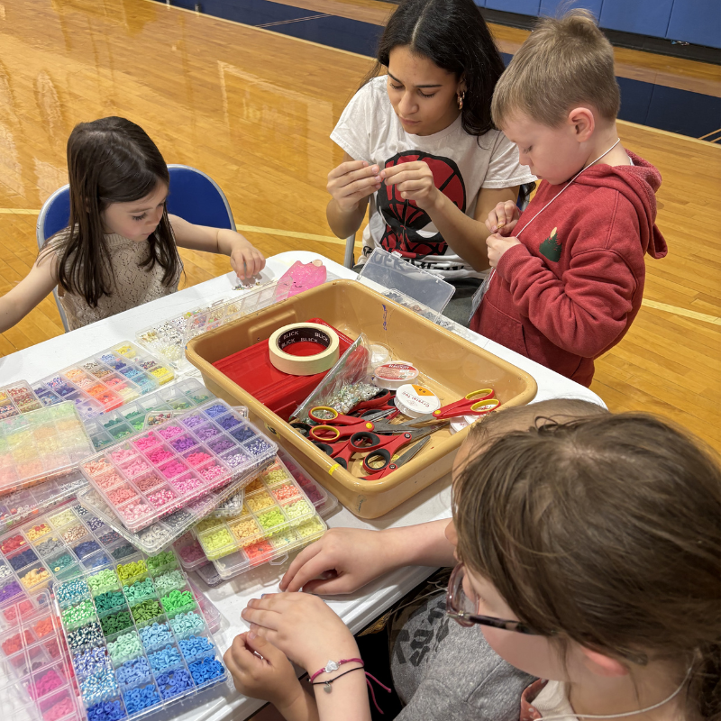 Children and adults are engaged in a beading activity, surrounded by colorful beads and craft supplies.