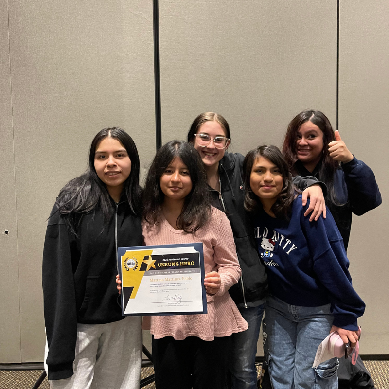 Five young women pose for a photo, with one holding a certificate for "UNSUNG HERO".