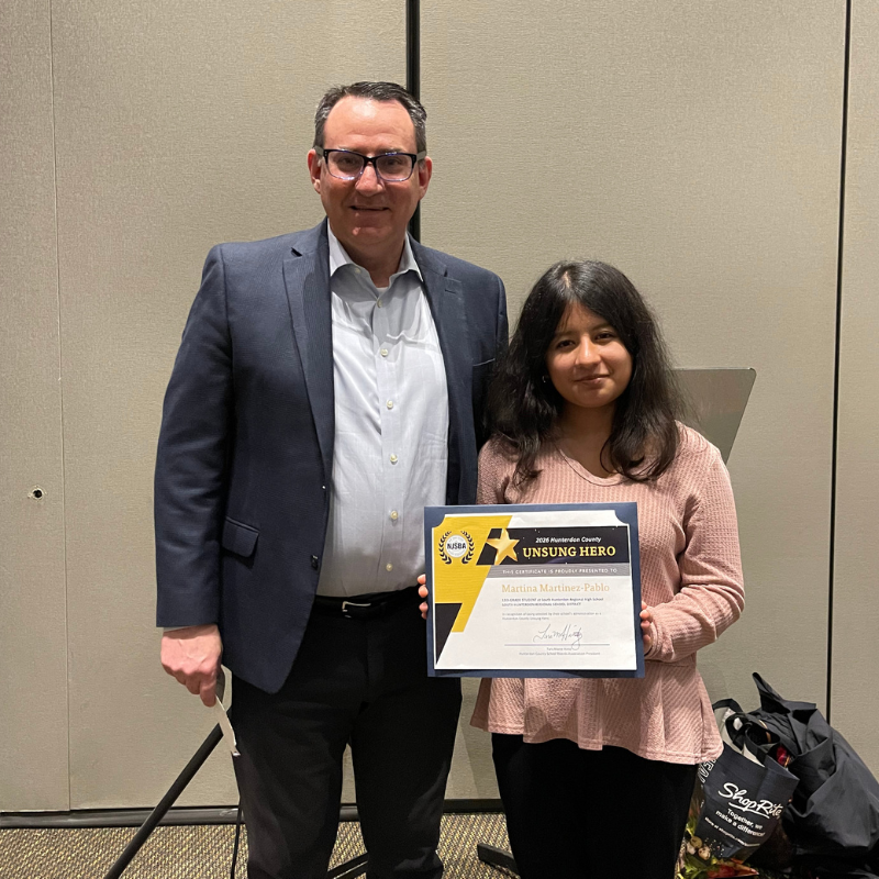 A man in a suit and a woman in a pink shirt stand together holding a certificate for "Unsung Hero".