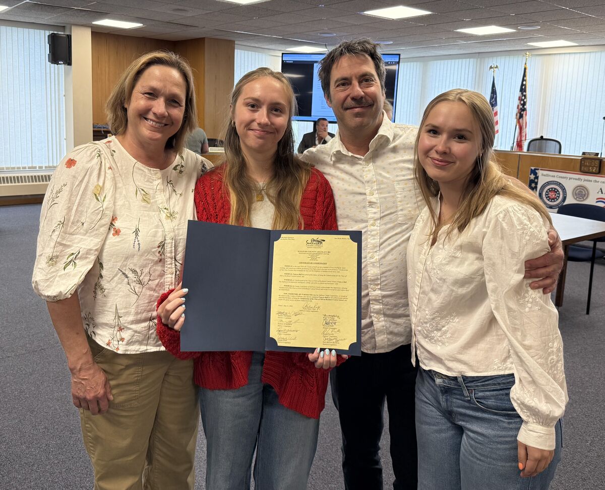 A family poses for a photo holding a certificate in a room with American flags and a screen.