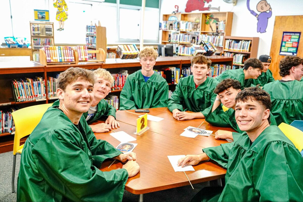 A group of smiling young men in green graduation gowns sit around a wooden table in a library.