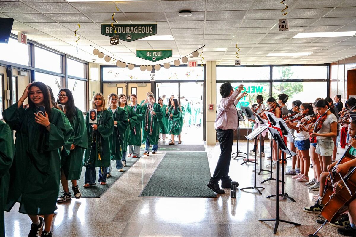 Students in green graduation gowns walk down a hallway, some holding photos, while an orchestra plays.