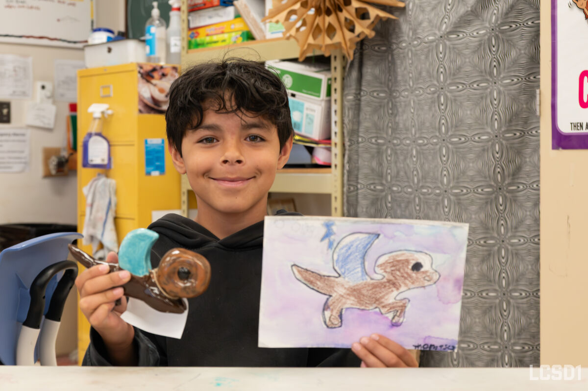 A young boy smiles while holding up a drawing of a winged creature and a clay sculpture.