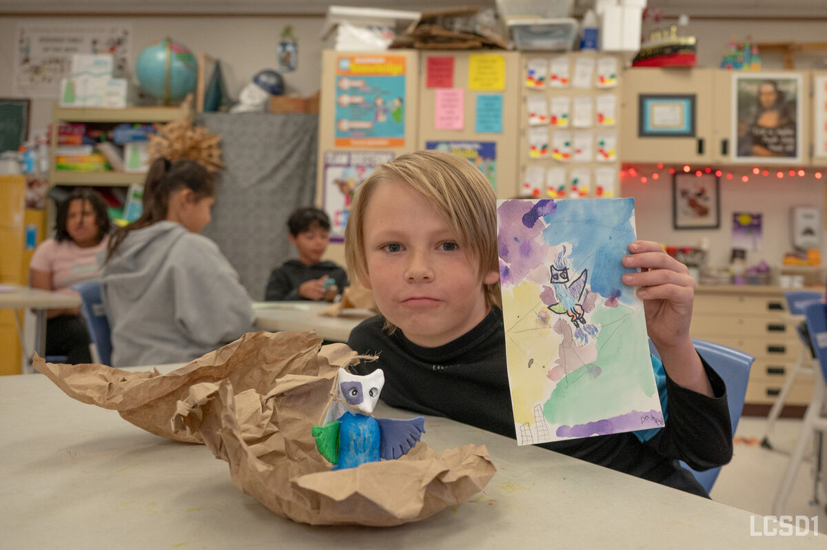 A young boy proudly displays his colorful watercolor painting and a small clay owl sculpture in an art classroom.