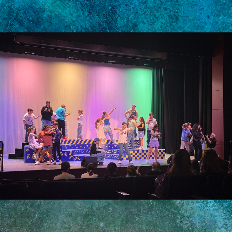 A group of young performers are on stage during a theatrical production, bathed in colorful stage lighting.