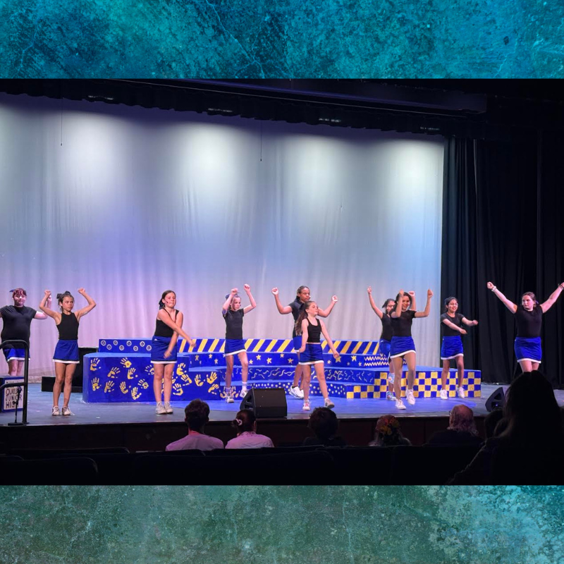 A group of young performers in black tops and blue skirts dance energetically on a stage.