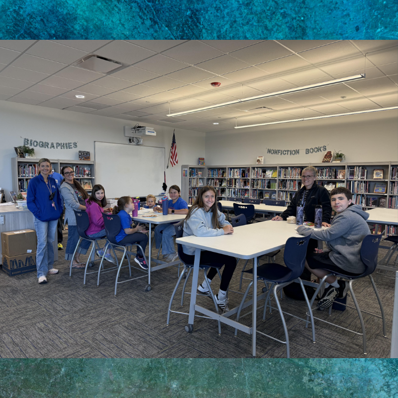 Students and adults gather around tables in a library with bookshelves in the background.