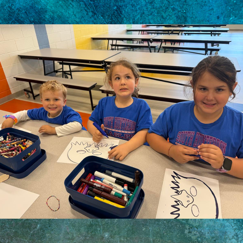 Three children in blue shirts sit at a table, drawing with crayons and markers.