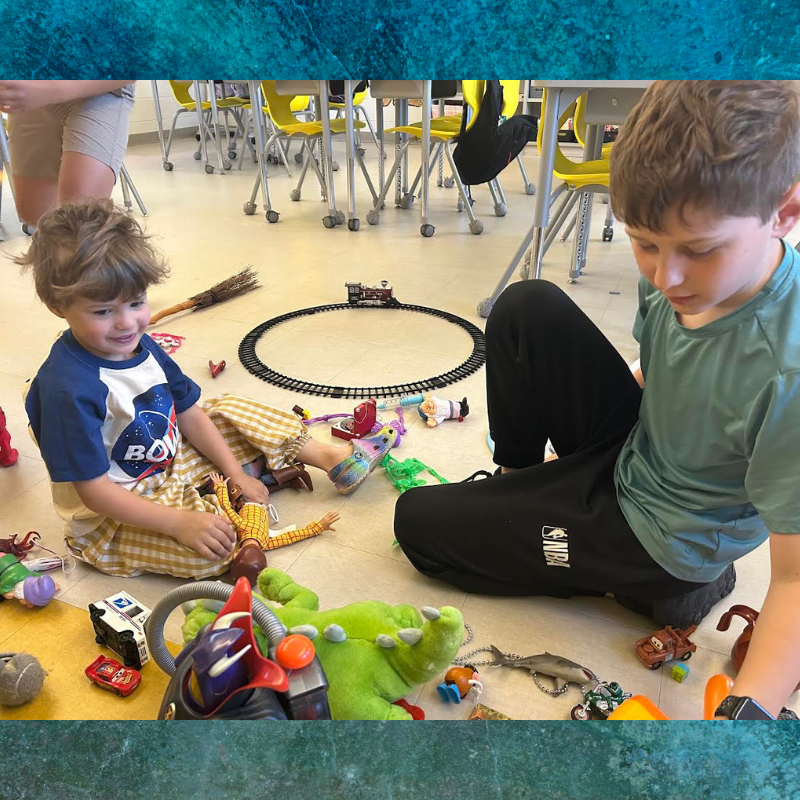 Two children play with a variety of toys, including a toy train set, on a classroom floor.