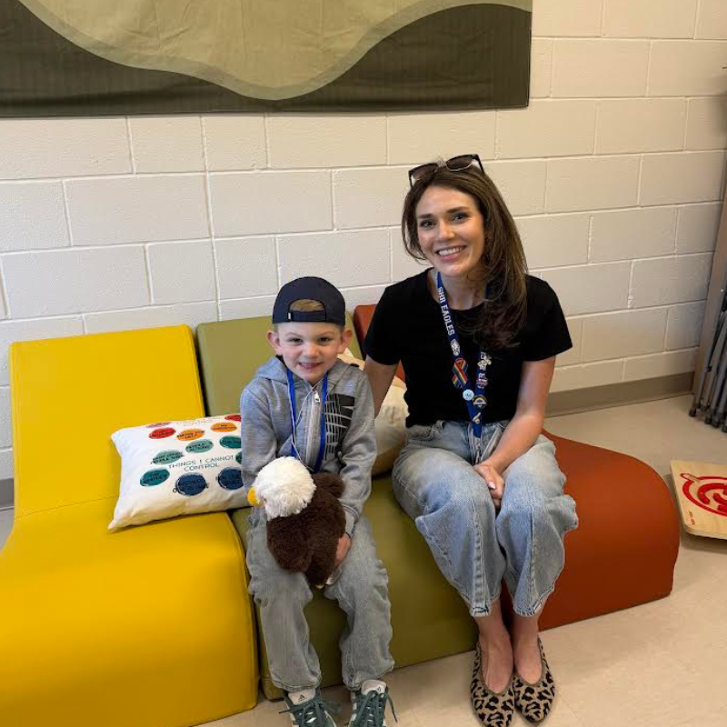 A smiling boy in a baseball cap holds a stuffed eagle while sitting next to a woman on colorful seating.