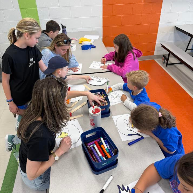 Children and adults are gathered around a table, drawing and coloring masks.