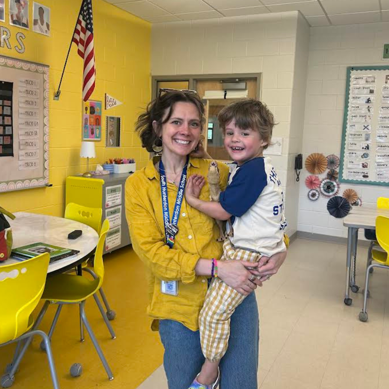 A smiling woman in a yellow shirt holds a young child in a classroom decorated with an American flag.