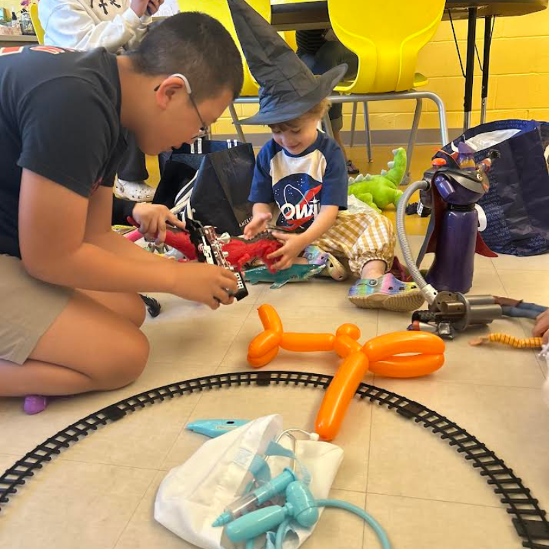 A young child wearing a witch hat plays with toys on the floor, surrounded by other children and toys.