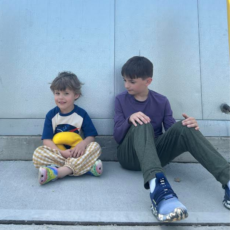 Two young boys sit on a concrete surface in front of a light blue wall.